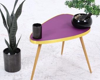 Könnte beinhalten: Kleiner Beistelltisch im Mid-Century-Stil mit lila Tischplatte und goldenem Rand. Der Tisch hat drei Holzbeine. Eine schwarze zylindrische Tasse und ein kleiner Bonsai-Baum in einem schwarzen Topf stehen auf dem Tisch. Links steht eine hohe, dunkelgrüne Pflanze in einem schwarzen Topf.