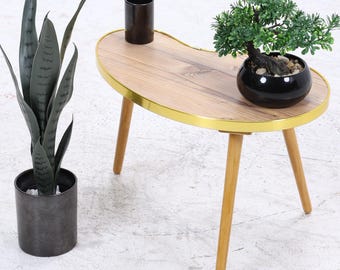 Könnte beinhalten: Kleiner, nierenförmiger Holztisch mit goldenem Rand und drei Holzbeinen. Auf dem Tisch stehen eine schwarze Topfpflanze und ein kleiner Bonsai in einem schwarzen Topf. Links steht eine hohe, dunkelgrüne Pflanze in einem dunkelgrauen Topf.