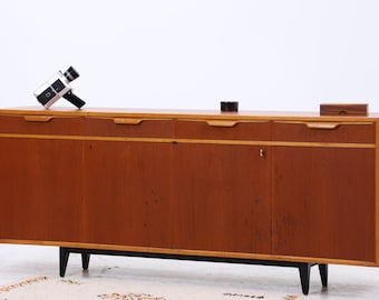 Könnte beinhalten: Ein Sideboard aus Holz im Mid-Century-Stil mit warmer brauner Oberfläche, drei Schubladen und zwei Türen. Der Schrank hat schwarze Beine und einen kleinen Teppich davor. Eine Vintage-Kamera und Dekorationsartikel befinden sich oben.