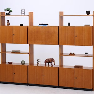 Op de afbeelding: Een houten roomdivider in mid-century stijl met meerdere planken en kasten. De unit is gemaakt van lichtgekleurd hout en heeft diverse decoratieve items, waaronder een bonsai boom, vazen en een houten olifantenbeeldje. Een vloerlamp met twee lampenkappen staat links.