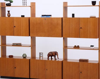 Könnte beinhalten: Ein Raumteiler aus Holz im Mid-Century-Stil mit mehreren Regalen und Schränken. Das Möbelstück ist aus hellfarbenem Holz gefertigt und verfügt über eine Vielzahl von Dekorationsgegenständen, darunter ein Bonsai-Baum, Vasen und eine Holz-Elefantenfigur. Eine Stehlampe mit zwei Lampenschirmen befindet sich links.