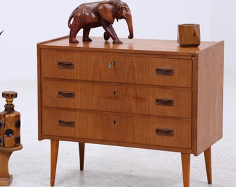 Könnte beinhalten: Mid-Century-Moderne Kommode aus Holz mit drei Schubladen, Schlüsselloch und Holzgriffen. Ein geschnitzter Elefant aus Holz und ein kleiner Holzblock sitzen oben. Eine Topfpflanze und eine dekorative Flasche befinden sich links.