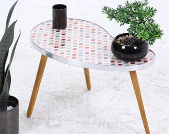 Könnte beinhalten: Ein Beistelltisch im Retro-Stil mit einem gemusterten Oberteil in Rot, Weiß und Blau. Der Tisch hat drei Holzbeine und einen Metallrand. Eine schwarze zylindrische Vase und eine kleine Topfpflanze stehen auf dem Tisch. Eine hohe, dunkelgrüne Pflanze steht in einem Metalltopf.
