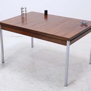 Könnte beinhalten: Ein rechteckiger Holztisch mit einer dunkelbraunen Oberfläche und silbernen Metallbeinen. Der Tisch hat ein schlichtes, modernes Design aus der Mitte des Jahrhunderts. Ein kleiner Aschenbecher und ein schwarzer Zylinder befinden sich auf dem Tisch. Ein dekorativer Pflanzenständer mit einer Pflanze und einer Flasche befindet sich links.