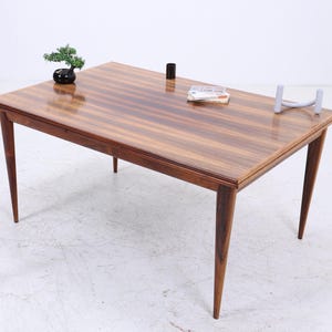 Puede incluir: Una mesa rectangular de madera con una superficie brillante y rayada. La mesa tiene cuatro patas cónicas y está adornada con pequeños objetos decorativos, incluyendo un bonsái, un cilindro negro y una pila de papeles. La mesa mide unos 152 cm de largo.