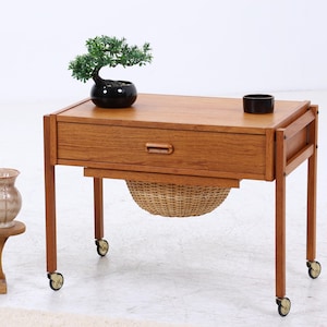 Könnte beinhalten: Ein Beistelltisch aus Holz im Mid-Century-Stil mit einer Schublade, einem Weidenkorb und Rollen. Obenauf steht ein kleiner Bonsai in einem schwarzen Topf sowie ein schwarzer Ring. Ein Pflanzenständer aus Holz hält eine Topfpflanze und eine dekorative Vase.