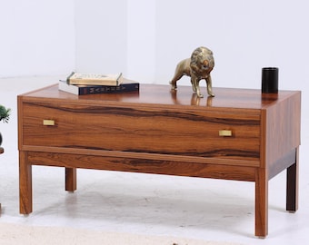 Könnte beinhalten: Ein Couchtisch aus dunklem Holz im Mid-Century-Modern-Stil mit zwei Schubladen und Messingbeschlägen. Auf der Oberseite befinden sich eine kleine Löwenstatue aus Messing, Bücher und ein schwarzer Zylinder. Ein Teppich und Pflanzen befinden sich im Vordergrund. Der Tisch ist ca. 120 cm lang.