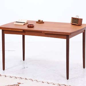 Pode incluir: Uma mesa de jantar de madeira de estilo moderno de meados do século com um tampo retangular e quatro pernas afiladas. A mesa é de cor castanha quente e tem uma extensão de folha. Um pequeno relógio, uma tigela e um livro estão sobre a mesa.