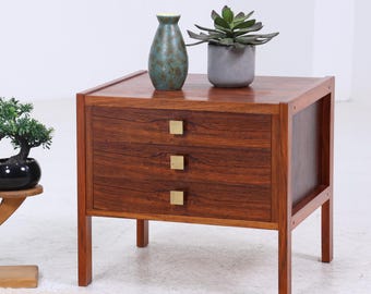 Könnte beinhalten: Ein Nachttisch aus Holz im Mid-Century-Modern-Stil mit drei Schubladen und Messingbeschlägen. Der Nachttisch ist mit einer grünen Vase, einer Sukkulente in einem grauen Topf und einem kleinen Pflanzenständer aus Holz geschmückt.