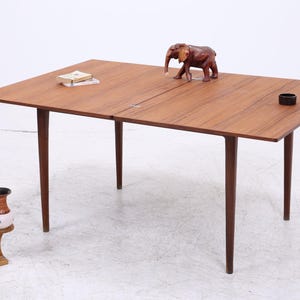 Pode incluir: Uma mesa de jantar retangular de madeira com quatro pernas cónicas, apresentando uma costura visível no centro. Na superfície da mesa encontram-se uma estatueta de elefante em madeira esculpida, um pequeno objeto circular preto e dois livros retangulares empilhados.