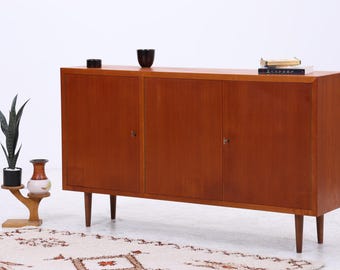 Könnte beinhalten: Ein rechteckiges Sideboard aus Holz mit drei Schranktüren und vier konischen Beinen. Obenauf stehen eine kleine schwarze Schale, ein dunkler Becher und ein Buch. Neben dem Sideboard steht ein Pflanzenständer aus Holz mit einer Pflanze.