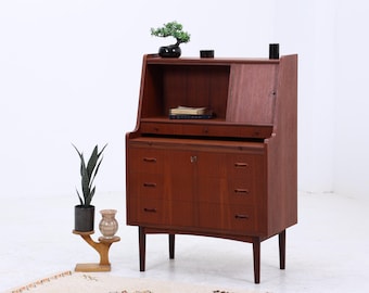 Könnte beinhalten: Ein Mid-Century-Schreibtisch aus Holz mit einer Hutze. Der Schreibtisch hat drei Schubladen und eine Schranktür. Ein kleiner Bonsai-Baum und andere dekorative Gegenstände stehen oben. Eine Topfpflanze und eine dekorative Vase stehen neben dem Schreibtisch.