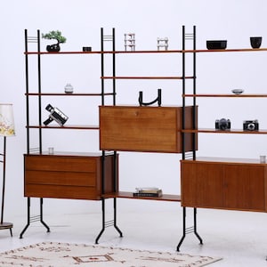 Peut inclure: Étagère de style moderne du milieu du siècle avec des supports en métal noir et des étagères, tiroirs et armoires en bois. L'unité est remplie d'objets décoratifs, dont un bonsaï, des appareils photo et de petites sculptures. Un lampadaire à deux abat-jours se trouve à gauche.