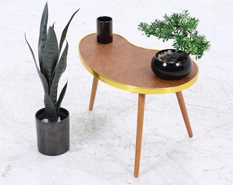 Könnte beinhalten: Ein Couchtisch im Mid-Century-Modern-Stil in Nierenform mit brauner Tischplatte, goldenem Rand und Holzbeinen. Auf dem Tisch stehen eine schwarze Topfpflanze und eine kleine schwarze Vase.
