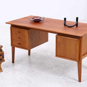 Op de afbeelding: Een houten bureau met een rechthoekig blad, drie lades aan de linkerkant en een enkele kastdeur aan de rechterkant. Het bureau heeft taps toelopende poten en wordt vergezeld door een kleine houten plantenstandaard met een plant en een glazen fles.
