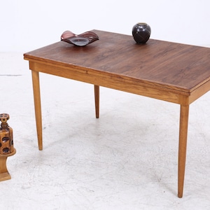 Op de afbeelding: Een rechthoekige houten eettafel met een donkerbruin blad en lichtbruine taps toelopende poten. Op de tafel staan een donkere glazen schaal en een kleine donkere vaas. Naast de tafel staat een houten standaard met een groene plant en een fles.
