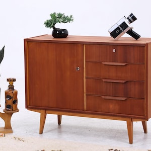 Könnte beinhalten: Ein hölzernes Sideboard mit einer Schranktür auf der linken Seite und vier übereinander angeordneten Schubladen auf der rechten Seite, die jeweils einen horizontalen Griff haben. Das Möbelstück steht auf vier konischen Beinen und hat ein Schlüsselloch.