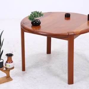 Peut inclure: Une table ronde en bois avec une finition lisse et polie et quatre pieds droits. Sur la surface de la table se trouvent une petite plante verte en pot et deux objets cylindriques sombres. À côté se trouve un petit support en bois avec une plante et un vase.