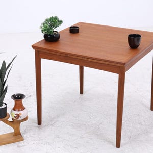 Peut inclure: Une table carrée en bois avec quatre pieds droits et une surface brune lisse. Sur la table se trouvent une petite plante verte en pot et deux bols sombres. À côté de la table se trouve un support en bois avec une grande plante et un vase.