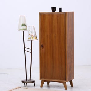 Pode incluir: Uma configuração de sala em estilo moderno de meados do século com um armário de madeira e uma luminária de chão. O armário é de cor castanho quente com uma única porta. A lâmpada tem dois abajures com desenhos florais.