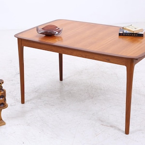 Peut inclure: Une table à manger rectangulaire en bois avec une surface brun clair lisse et une bordure brun foncé. La table possède quatre pieds fuselés. Sur la surface se trouvent un bol en verre foncé et un livre intitulé Mastery. Un petit support en bois porte une plante serpent et une carafe en verre brun.