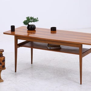 Peut inclure: Une table basse de style moderne du milieu du siècle avec un plateau rectangulaire et une étagère inférieure. La table est en bois avec une finition marron chaud. Un petit bonsaï et deux objets cylindriques noirs sont posés sur la table.