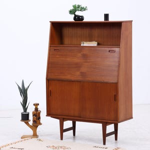 Puede incluir: Un escritorio secreter alto de madera de teca con un estante superior abierto, una superficie de escritura central abatible y un armario inferior con dos puertas correderas. La unidad se apoya en cuatro patas cónicas junto a un bonsái y una jarra.