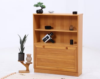 Könnte beinhalten: Ein hellbraunes Bücherregal aus Holz mit zwei Regalen und einem Unterschrank. Auf den Regalen stehen ein kleiner Bonsai-Baum, eine Vintage-Kamera und verschiedene Dekorationsartikel. Eine hohe, dunkelgrüne Pflanze und eine kleine Vase stehen auf einem Holzständer.