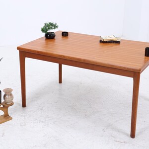 Op de afbeelding: Een rechthoekige houten tafel met vier poten, afgewerkt in lichtbruin. Het gladde oppervlak is versierd met kleine decoratieve items, waaronder een plant, een boek en twee zwarte cilindrische objecten. De tafel is ongeveer 152 cm lang.