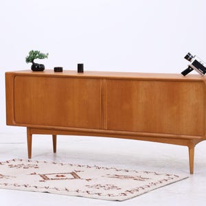 Può includere: Una credenza in legno color marrone chiaro con due ante scorrevoli e gambe affusolate. Sopra sono posizionati un piccolo bonsai, due oggetti cilindrici neri e una cinepresa vintage. Accanto si trova un piccolo supporto in legno con pianta e vaso.