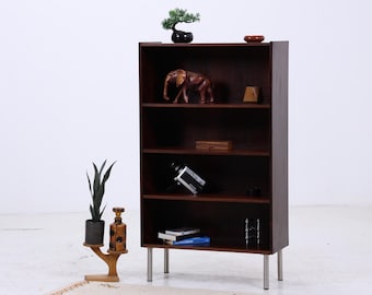 Könnte beinhalten: Ein dunkelbraunes Bücherregal aus Holz mit vier Regalen, getragen von silbernen Beinen. Die Regale zeigen dekorative Gegenstände, darunter einen Bonsai-Baum, eine Elefantenfigur aus Holz und eine kleine Holzkiste. Eine Topfpflanze und eine dekorative Flasche stehen auf einem Beistelltisch.