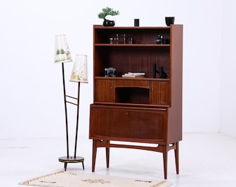 May include: A mid-century modern wooden cabinet with a hutch and a lower cabinet. A two-bulb floor lamp with patterned shades stands to the left. The cabinet has shelves with decorative items. A small rug is on the floor.
