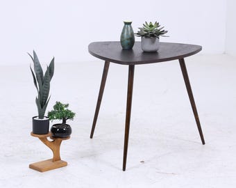 Könnte beinhalten: Ein dunkelbrauner Beistelltisch im Mid-Century-Modern-Stil mit dreieckiger Form und drei abgewinkelten Beinen. Auf dem Tisch stehen eine grüne Vase und eine Topfpflanze. Ein Pflanzenständer aus Holz zeigt zwei Topfpflanzen.