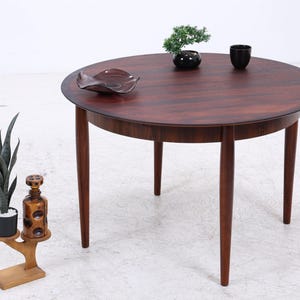 Peut inclure: Une table à manger ronde en bois brun foncé avec quatre pieds fuselés. Sur la surface de la table se trouvent un petit bonsaï vert dans un pot noir, une tasse noire et un plat ondulé brun foncé. À côté, un support en bois clair porte une plante et une bouteille.