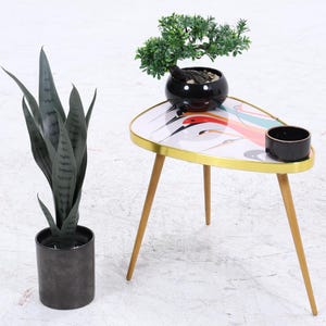 Op de afbeelding: Een bijzettafel in retrostijl met een kleurrijk abstract ontwerp op het tafelblad. De tafel heeft een gouden rand, drie houten poten en een kleine zwarte keramische kom. Een pot met slangplant en een bonsai zijn ook aanwezig.