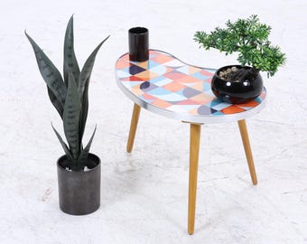 Könnte beinhalten: Kleiner, nierenförmiger Beistelltisch mit farbenfrohem geometrischem Muster auf der Tischplatte. Der Tisch hat drei Holzbeine und eine silberne Umrandung. Eine schwarze zylindrische Vase und eine kleine Topfpflanze stehen auf dem Tisch. Links steht eine hohe, dunkelgrüne Pflanze in einem schwarzen Topf.