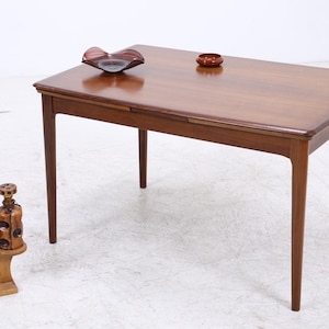Peut inclure: Une table à manger rectangulaire en bois avec des pieds fuselés et une finition lisse. Sur la table se trouvent un bol en verre rouge foncé et un petit plat marron. À côté, un support en bois porte une plante verte et une bouteille en verre marron.