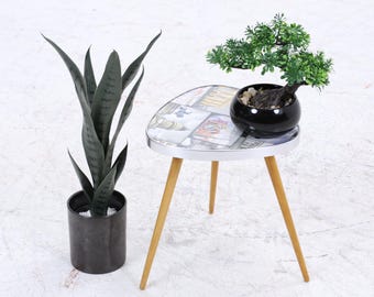 Könnte beinhalten: Ein kleiner, runder Beistelltisch mit weißer Tischplatte und Holzbeinen. Auf dem Tisch steht ein kleiner Bonsai-Baum in einem schwarzen Topf und eine dekorative Pflanze. Links steht eine hohe, dunkelgrüne Pflanze in einem schwarzen Topf.