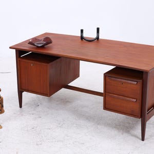 Op de afbeelding: Een rechthoekig houten bureau met een warme bruine afwerking, voorzien van een kast aan de linkerkant en een unit met twee lades aan de rechterkant. Het bureau heeft taps toelopende poten en een vlak bovenblad met een plant en een bruine fles.