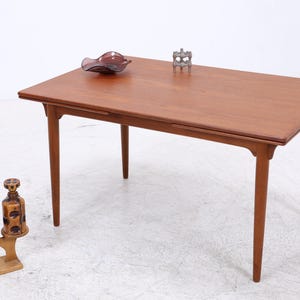 Pode incluir: Uma mesa de jantar retangular em teca com pernas cónicas e uma superfície de madeira lisa. Sobre a mesa, encontra-se uma taça de vidro roxo escuro e um pequeno castiçal de metal cinzento. Ao lado da mesa, um suporte de madeira sustenta uma planta.
