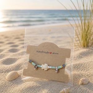 May include: A handmade bracelet featuring a sea turtle charm, shell charms, and blue and white beads. The bracelet is presented on a sandy beach with seashells. The packaging reads "Handmade with love".