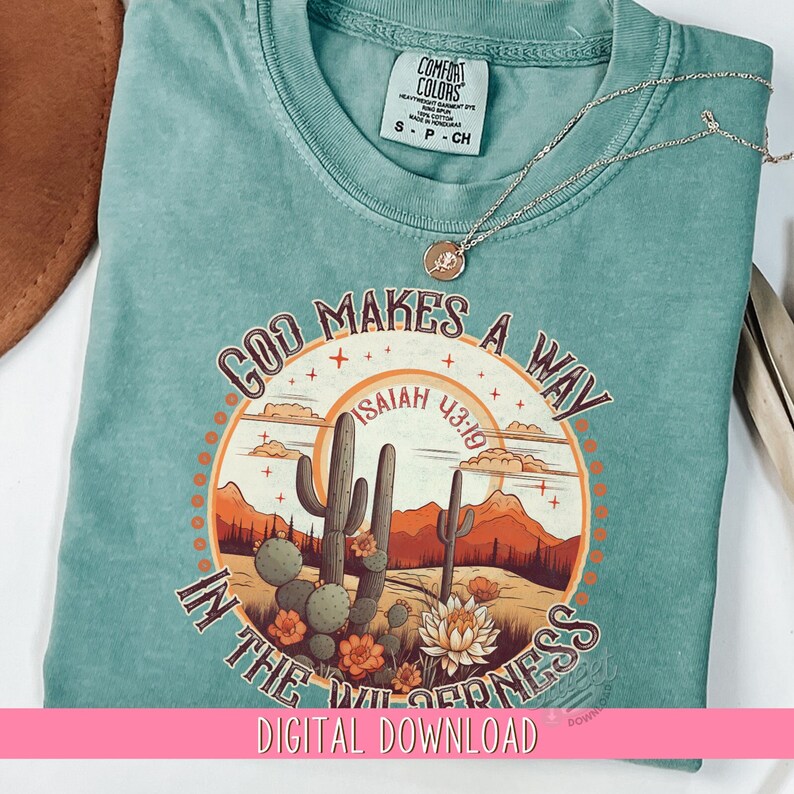 May include: A teal blue t-shirt with a graphic design of a desert scene with cacti and mountains. The graphic includes the text "God Makes a Way" and "Isaiah 43:19".