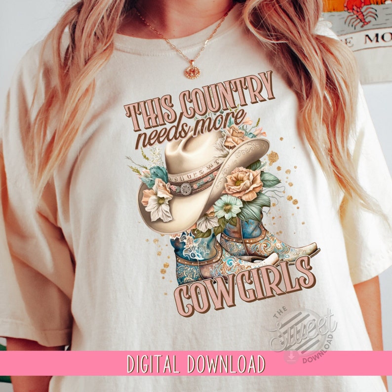 May include: A white t-shirt with the text "This country needs more cowgirls" in pink lettering. The design features a cowboy hat with floral accents and two pairs of cowboy boots with floral patterns.