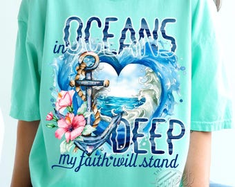 Puede incluir: Una camiseta azul turquesa con un diseño de acuarela de un ancla y olas en forma de corazón. El texto "in oceans deep my faith will stand" está impreso en la camiseta.