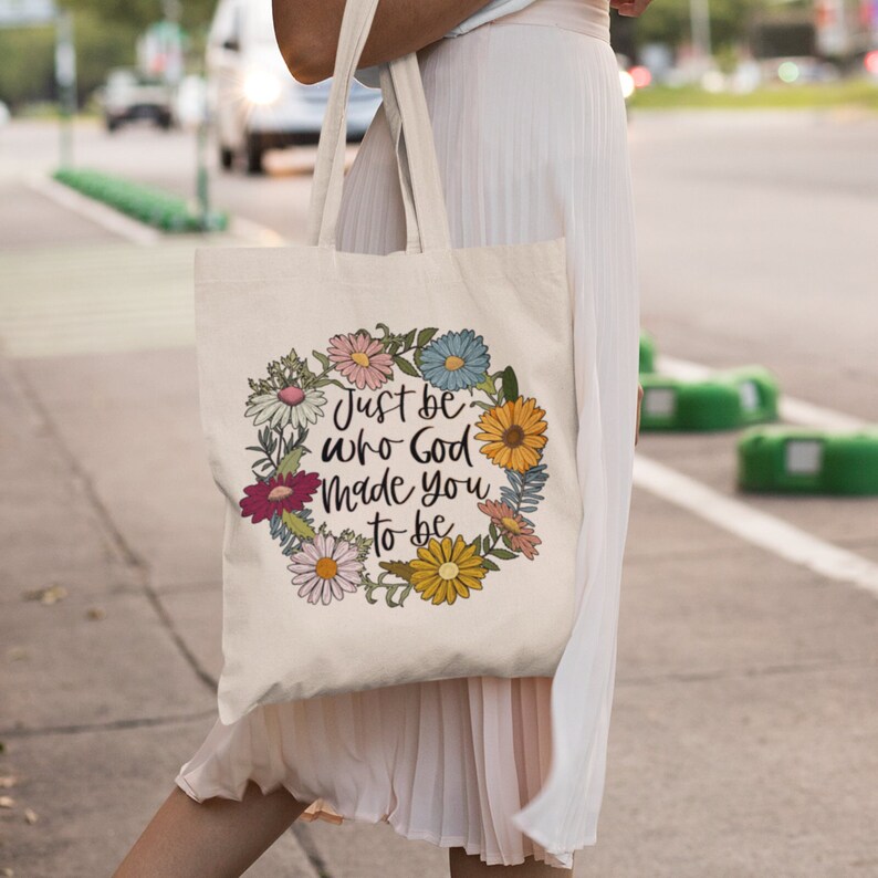 May include: A white canvas tote bag with a floral wreath design and the text "Just be who God made you to be".