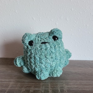 May include: A handmade, teal-colored crochet frog plushie with black safety eyes and a stitched mouth. The frog has a textured, fluffy appearance and small arms and legs. The toy is sitting on a wooden surface.