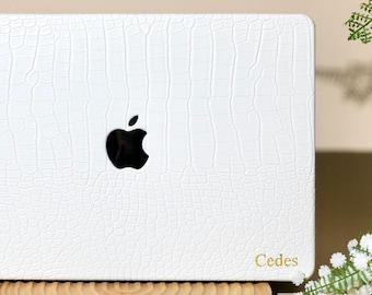 May include: A white laptop cover with a textured crocodile skin pattern, featuring a black Apple logo in the center and the name Cedes printed in gold in the bottom right corner. The cover is positioned on a circular wooden stand.