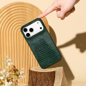 Könnte beinhalten: Eine dunkelgrüne, strukturierte Handyhülle mit Kameraausschnitt, die auf einem kleinen Holzstumpf liegt. Die Hülle ist der Fokus, mit einer Hand, die darauf zeigt. Der Hintergrund umfasst einen Holzbogen, Blumen und den Text "Ninetydegree."