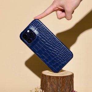 May include: A blue crocodile-embossed phone case on a wooden stump with moss and flowers. The case is positioned at a 90-degree angle.