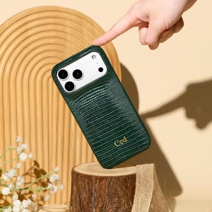 Op de afbeelding: Een donkergroen smartphonehoesje met een textuur van hagedissenhuid en de in goud gedrukte letters Ced. Het hoesje heeft een rechthoekige uitsparing voor de camera met drie ronde lenzen en een flitser.